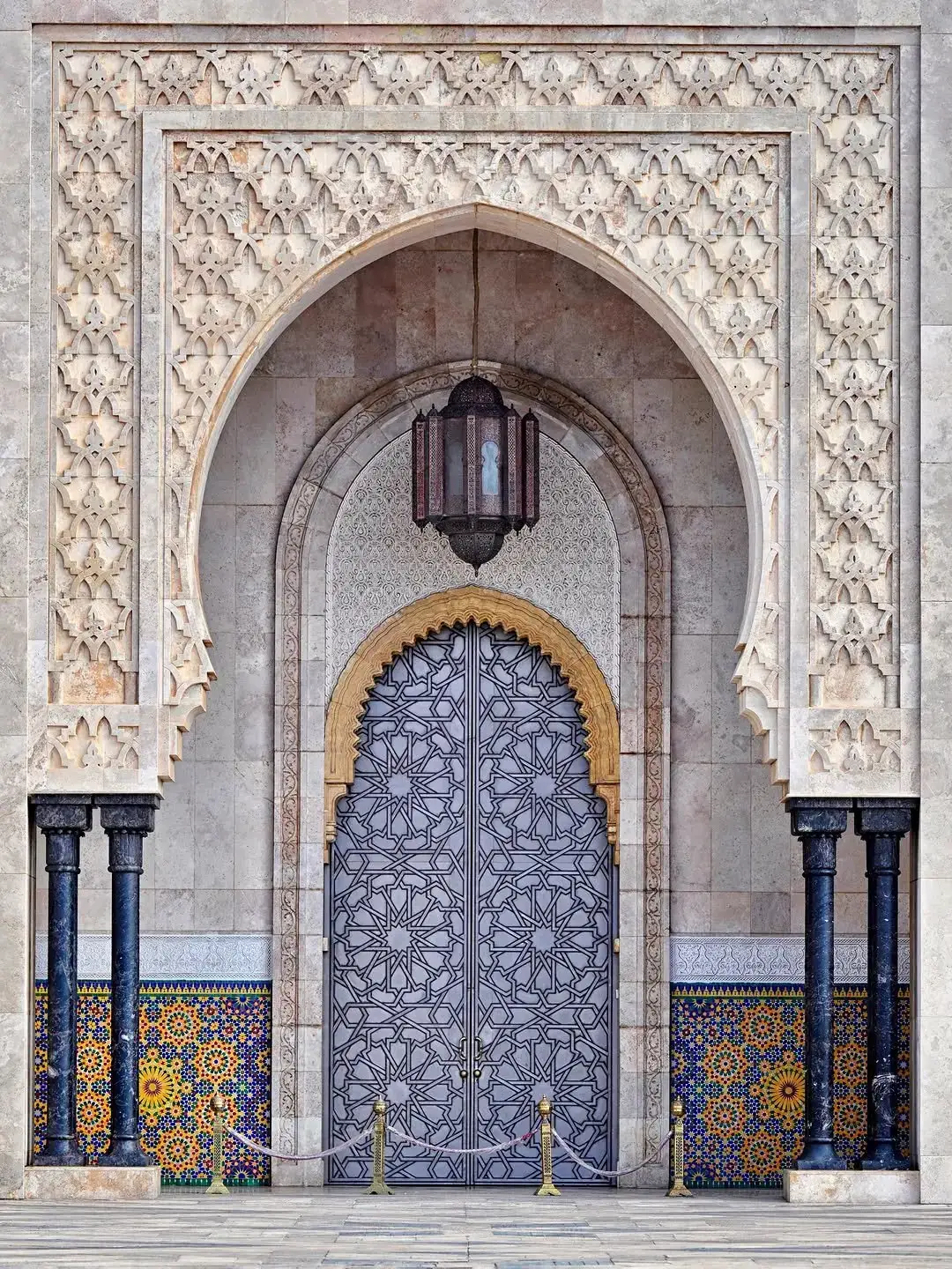 Ornate pointed-arch doorway of the Hassan II Mosque in Casablanca — carved marble arabesques, geometric metalwork doors and zellige tilework