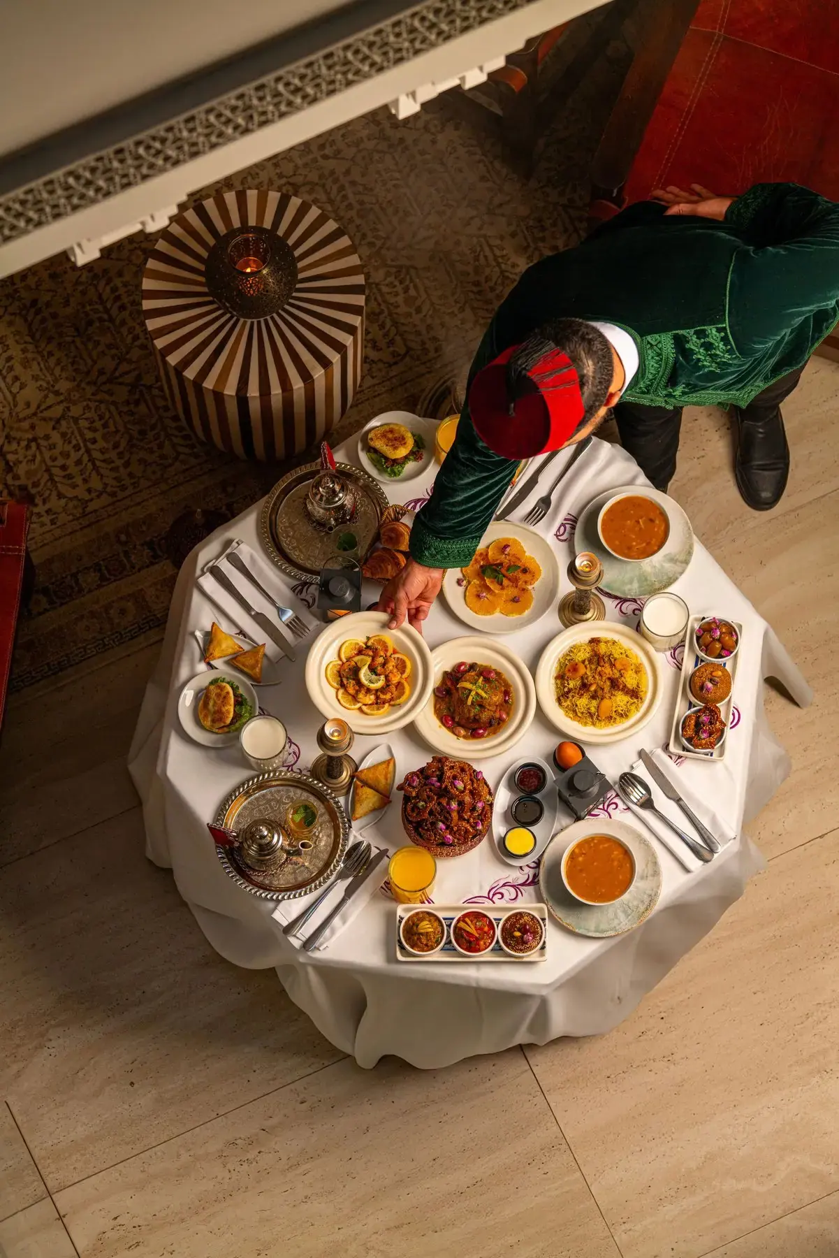 Waiter in a green djellaba and red tarbouche presenting a full spread of Moroccan dishes on a round riad table
