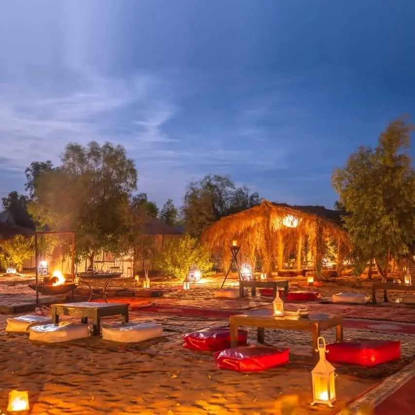 Saharan bivouac lounge with lanterns, carpets and a thatched canopy at dusk near Merzouga