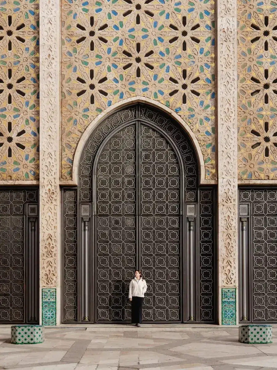 Woman standing before the giant ornate bronze door and mosaic wall of the Hassan II Mosque, Casablanca