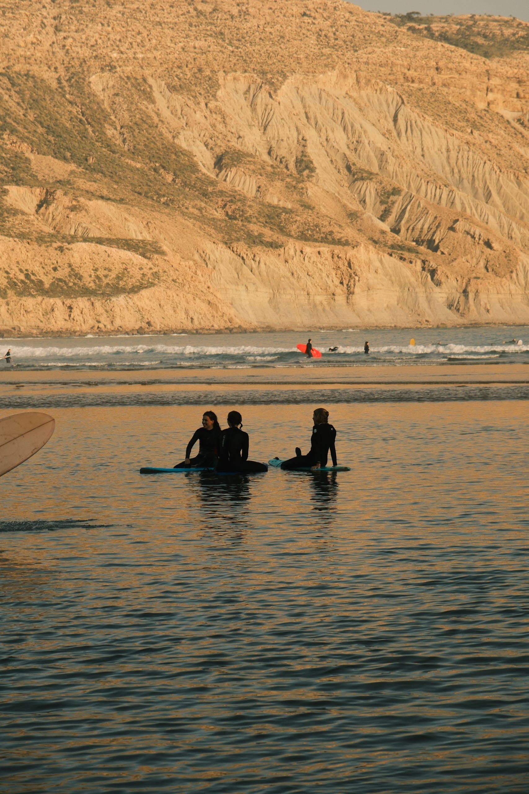 Three surfers on boards at golden hour with a Moroccan rocky cliff coast behind