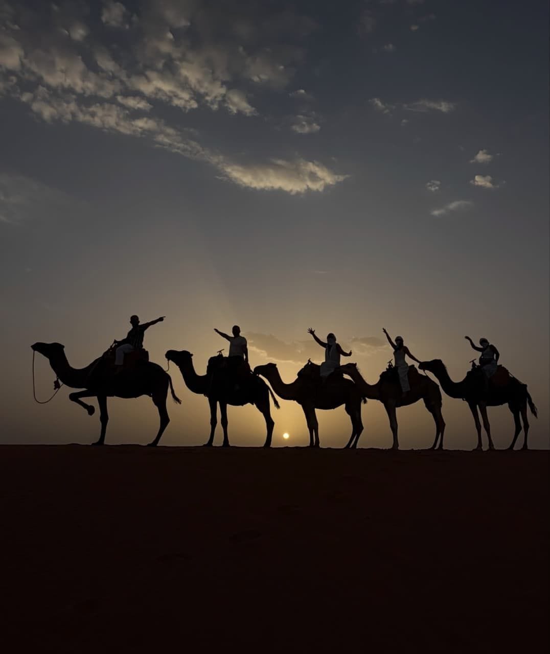 Riders waving from a line of camels silhouetted against a low sun