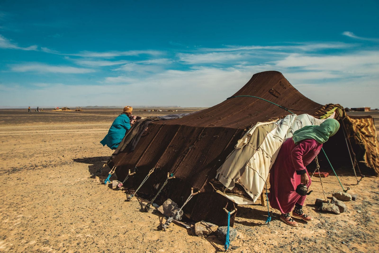 A woven nomad tent on open, stony desert ground, with two people in long robes beside it