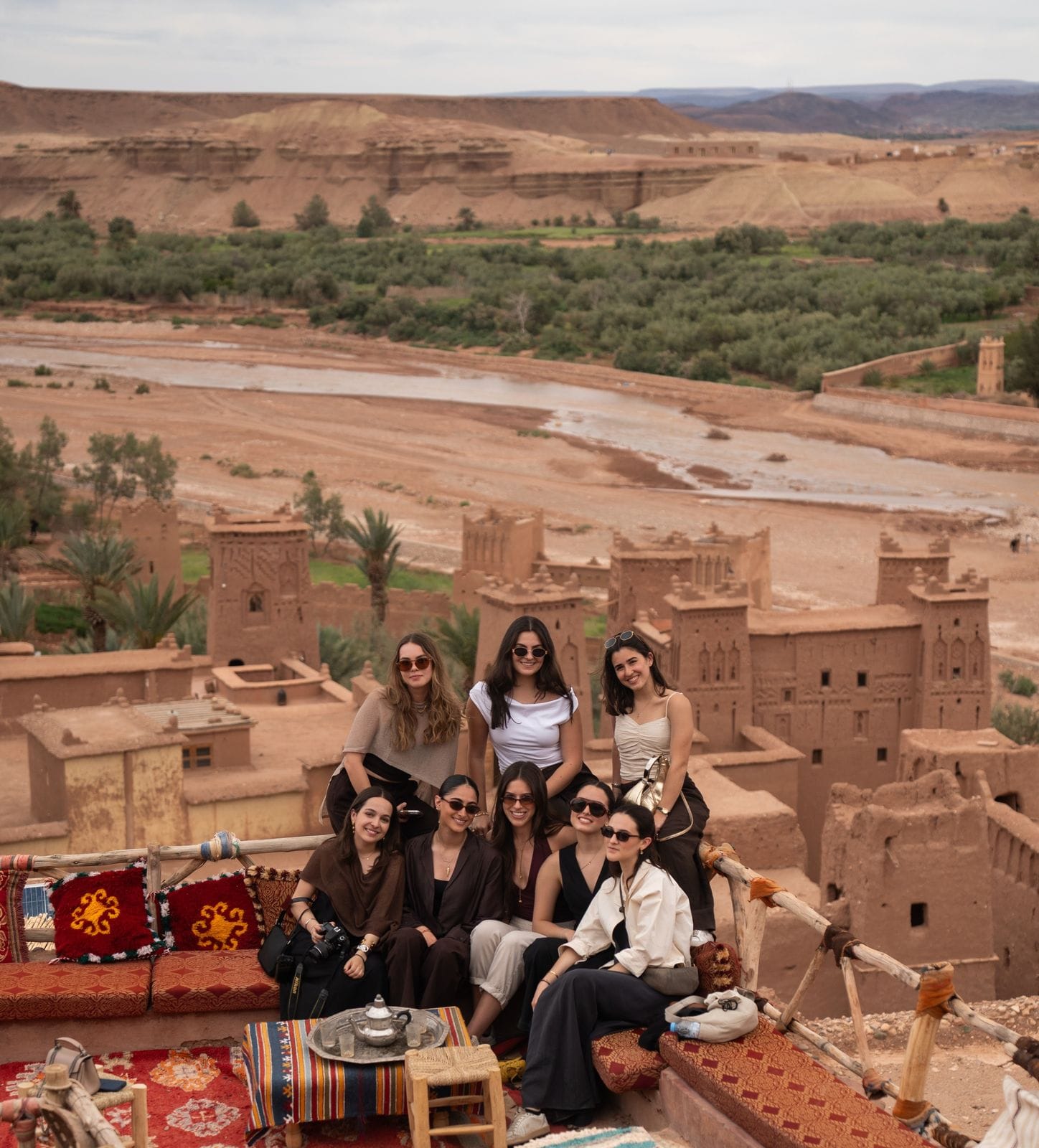 Students on a rooftop terrace overlooking earthen kasbahs, a riverbed and a palm grove