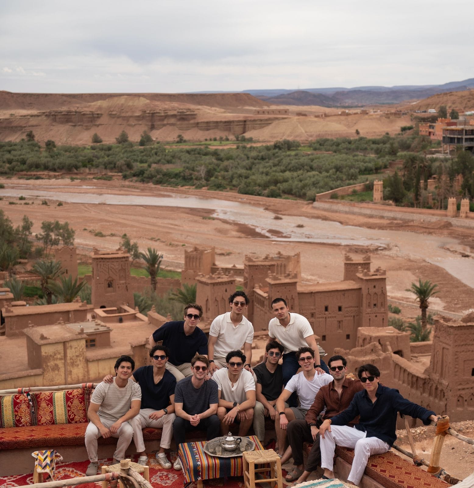 Students on a rooftop terrace above earthen kasbahs, a dry riverbed and palm trees